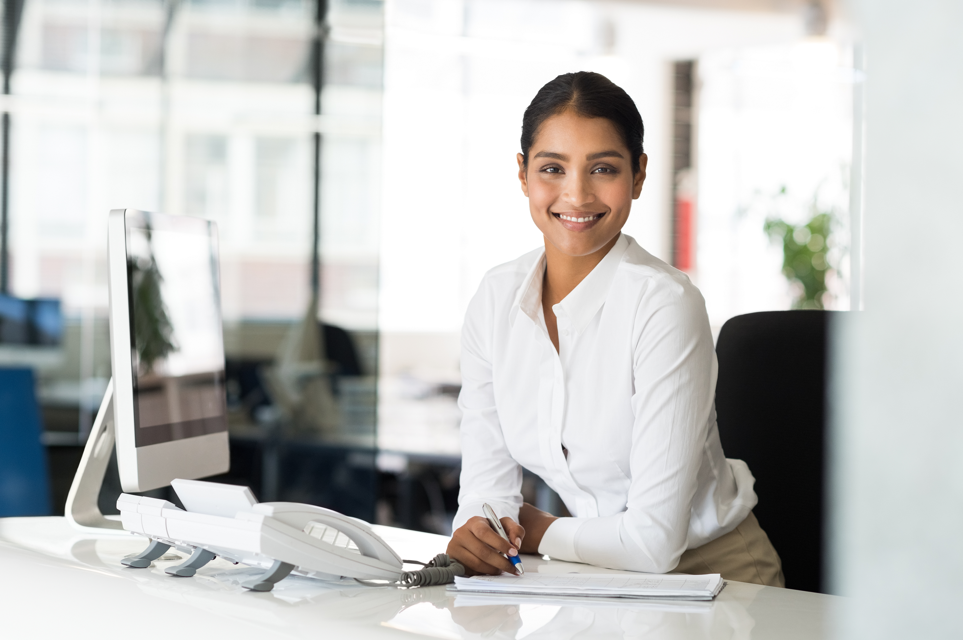Mujer sonriente, frente a escritorio y computadora en oficina amplia. ECE. Trimestre DEF 2023-2024.
