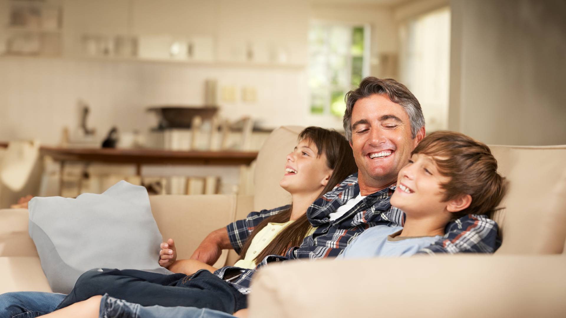 Padre en sofá, sonriendo en compañía de hijo e hija infantiles. Especial Día del Padre 2024.