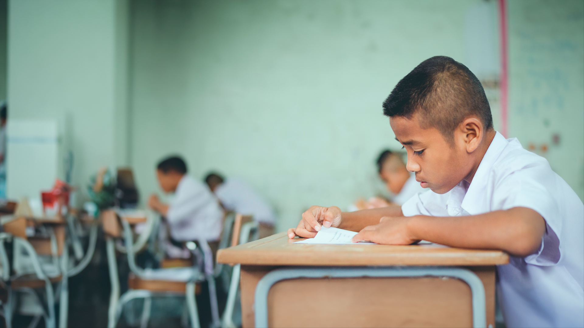 Niño escolar sentado junto a su pupitre en el aula observa hoja de papel. ENAHO 2023. Infografía especial de educación.