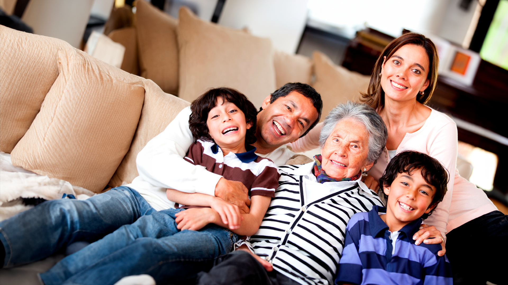 Familia con papá, mamá y abuela y dos hijos se abrazan sentados en sofá mientras sonríen. Día de la Madre 2024.