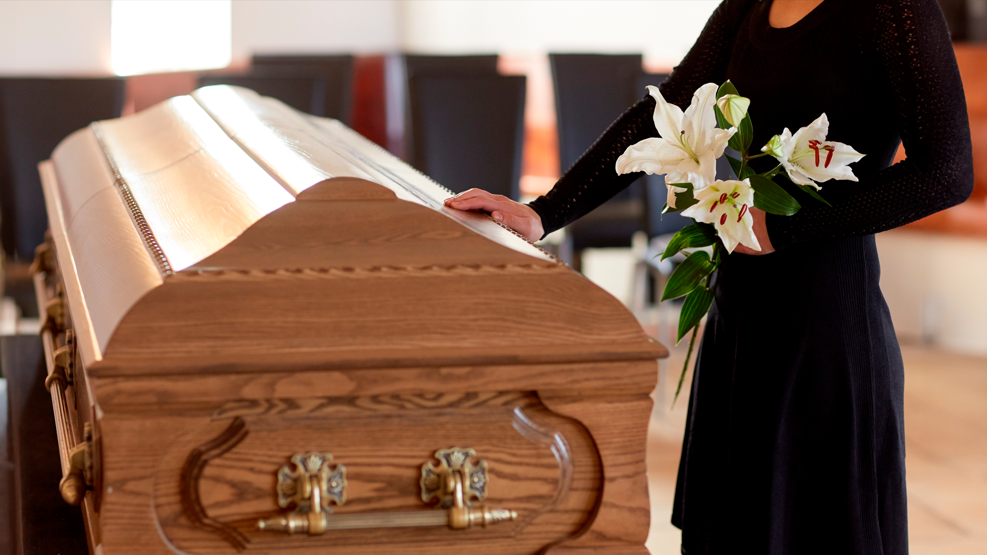 Mujer con vestido negro sostiene ramos de flores y posa su mano sobre ataúd en sala funeraria. Especial del Día de los Fieles Difuntos 2024.