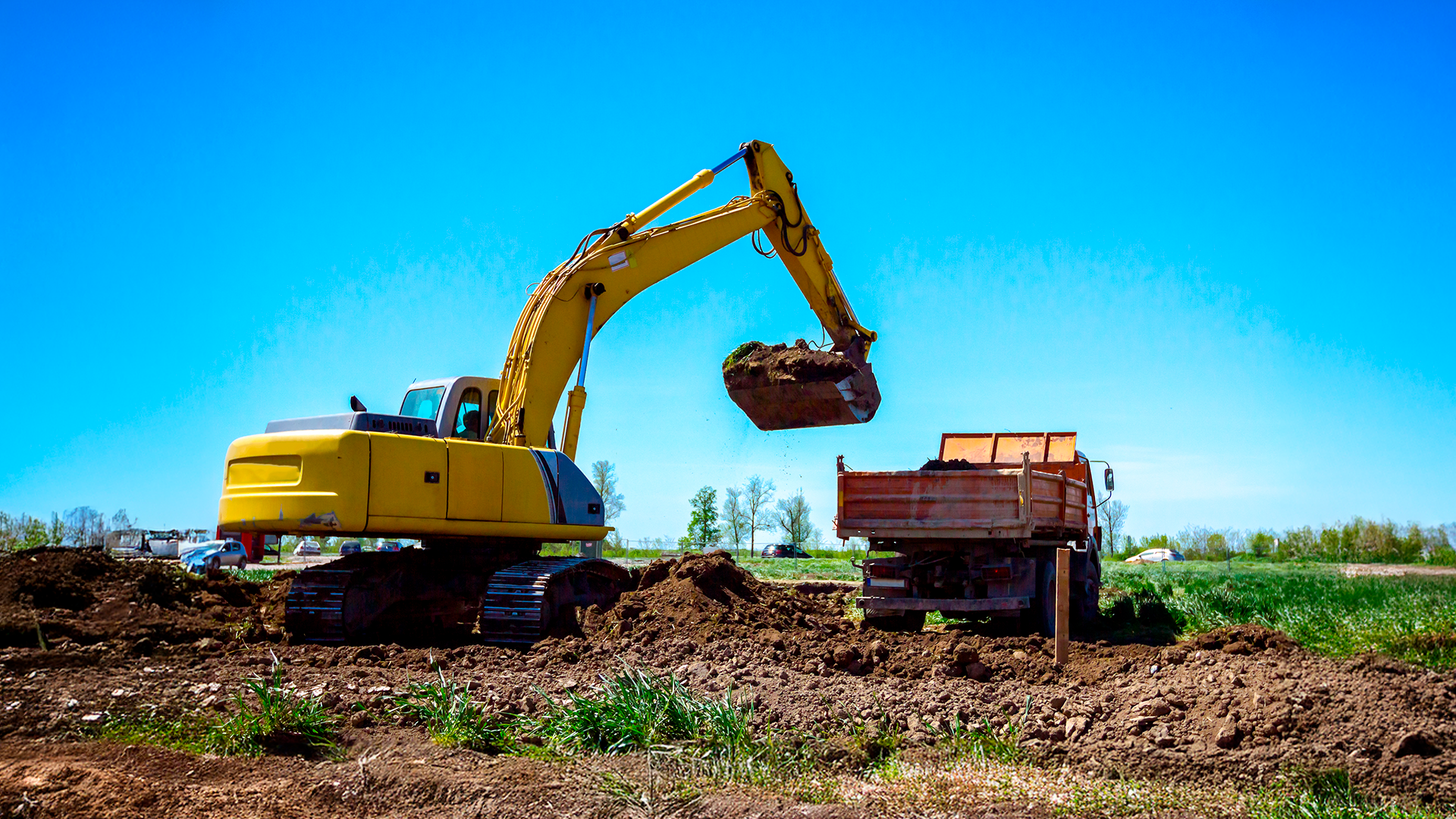 Escavadora mecánica carga tierra en cajón de vagoneta durante movimiento de tierras para obra de construcción. IPCons. FEB 2025.