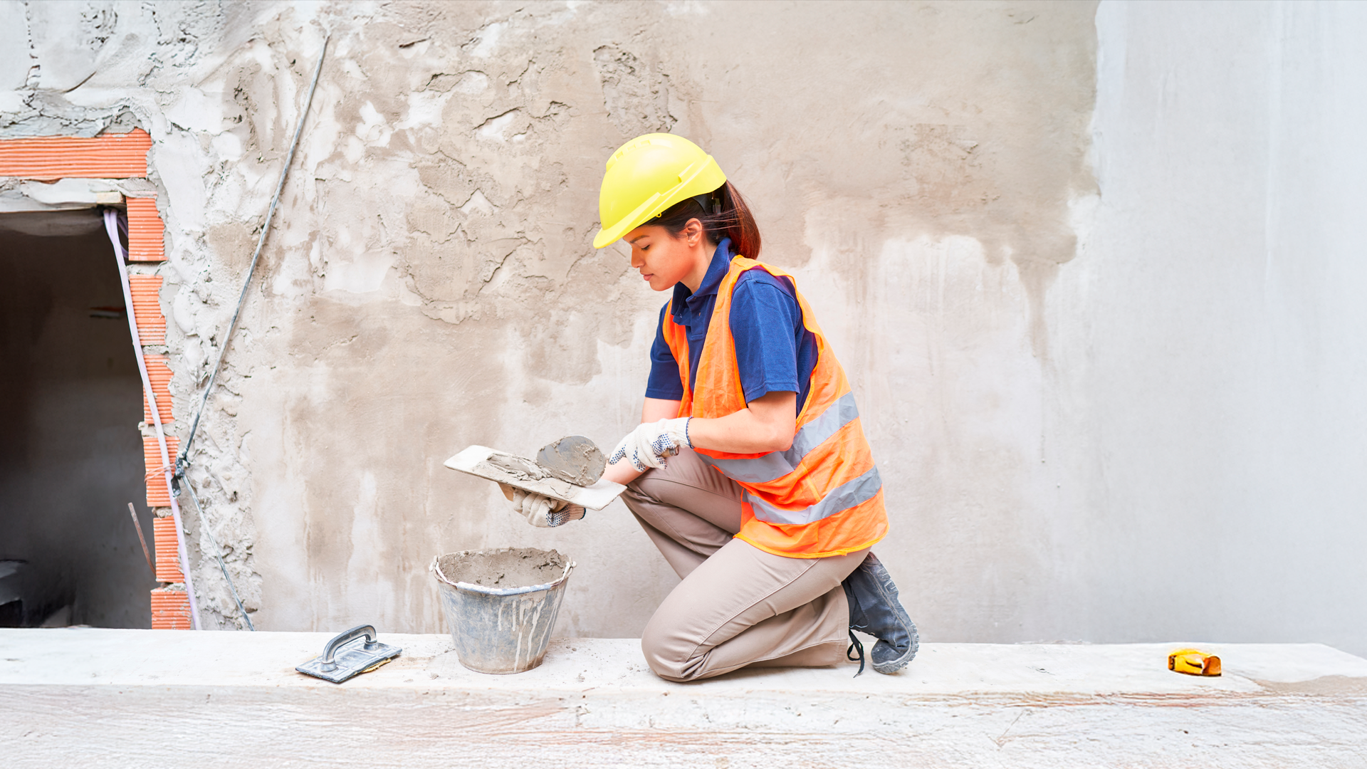 Mujer trabajadora de la construcción prepara mezcla de concreto para repello de pared. IPCons. ABR 2025.
