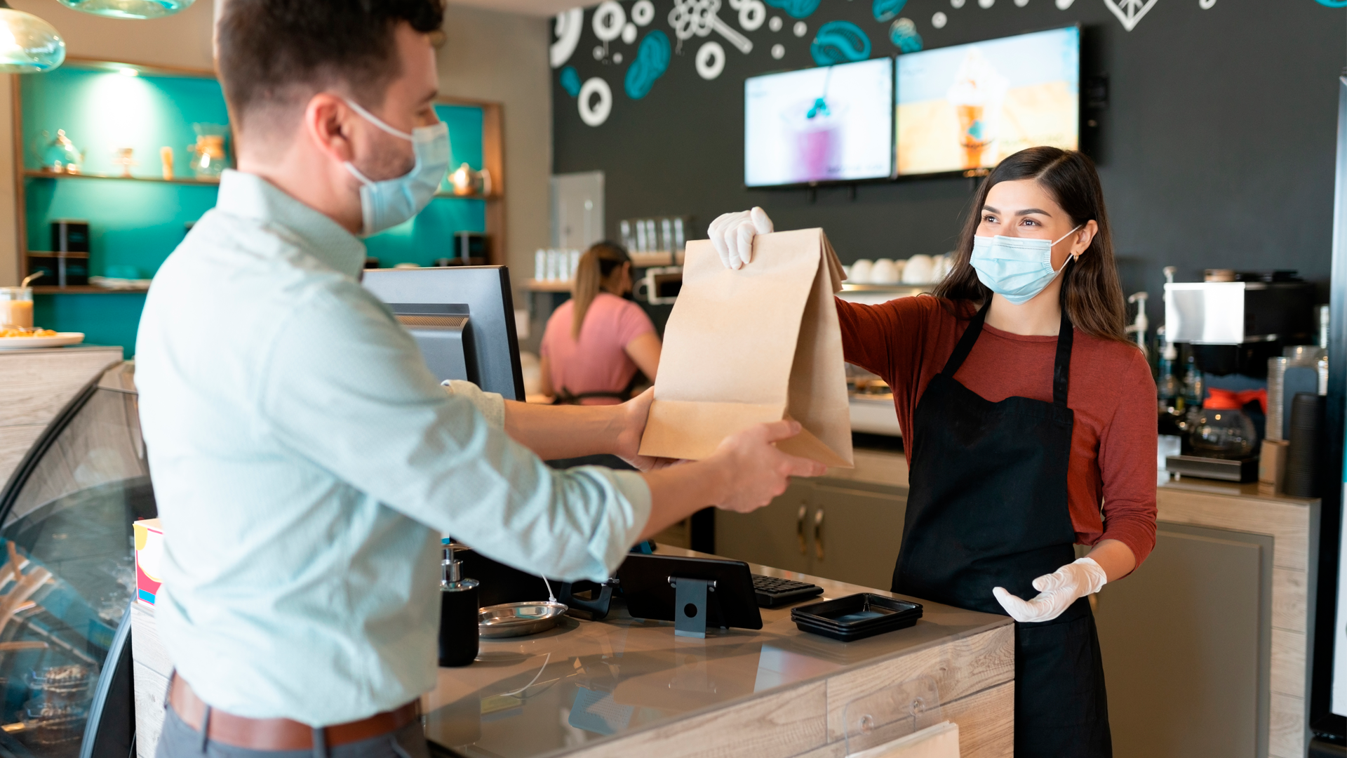 Mujer entrega bolsa de papel a cliente frente a mostrador de cafetería. DEE 2022.