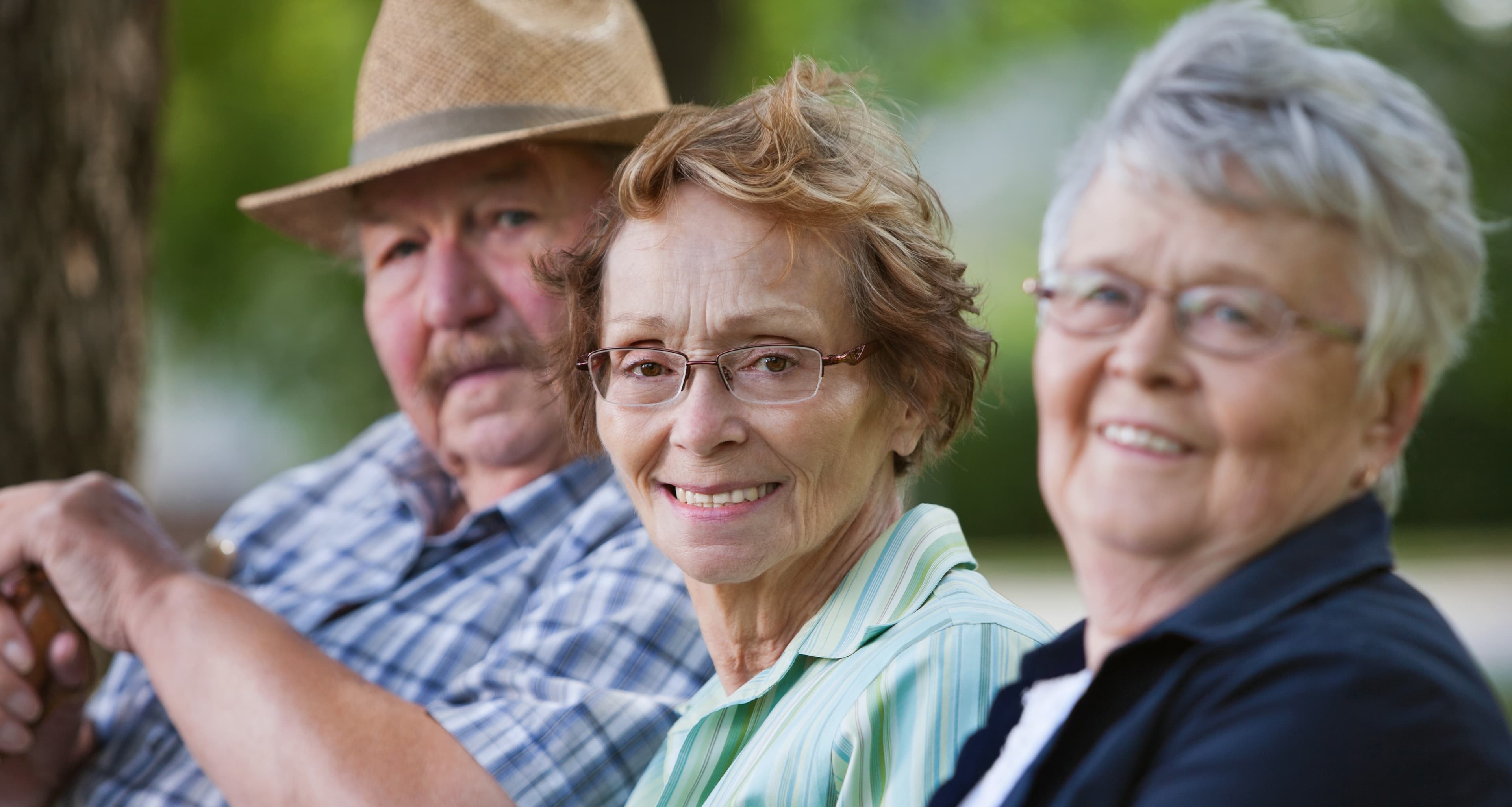 Tres personas mayores sentadas en la banca de un parque sonríen viendo hacia la cámara. Día de las Personas Mayores 2023.