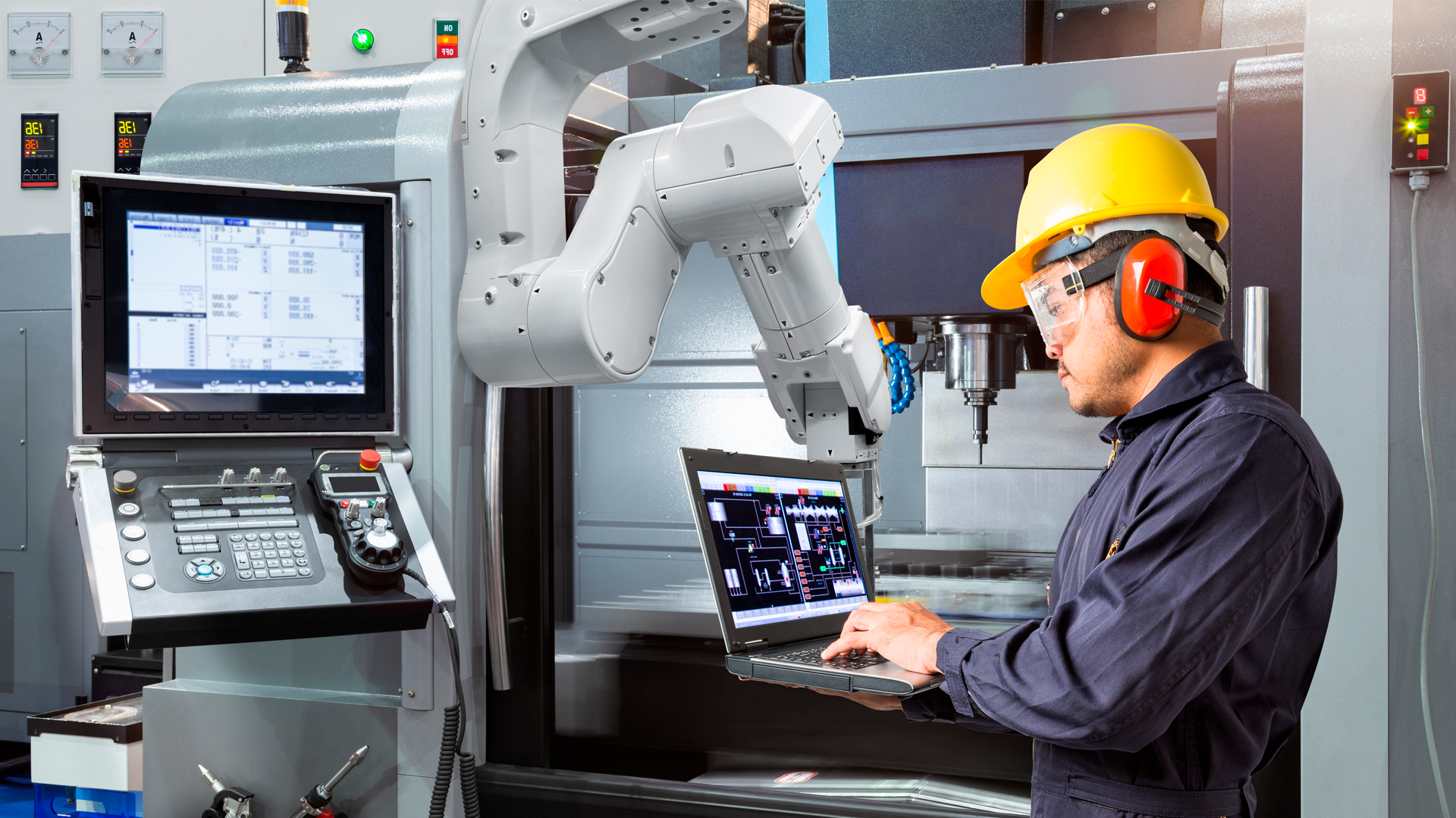 Trabajador con casco y audífonos de protección utiliza computadora portatil mientras revisa robot en planta de ensamblaje. ECE, Trimestre JJA 2023.